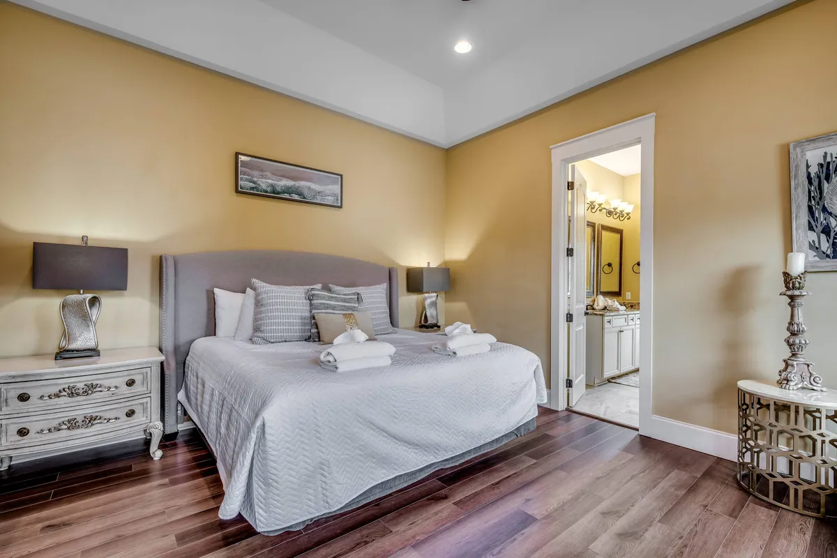 A tranquil bedroom featuring a neatly made bed, nightstands with lamps, tasteful artwork above the headboard, and a visible doorway that leads to a bathroom. The space feels cozy and private, emphasizing relaxation and comfort.