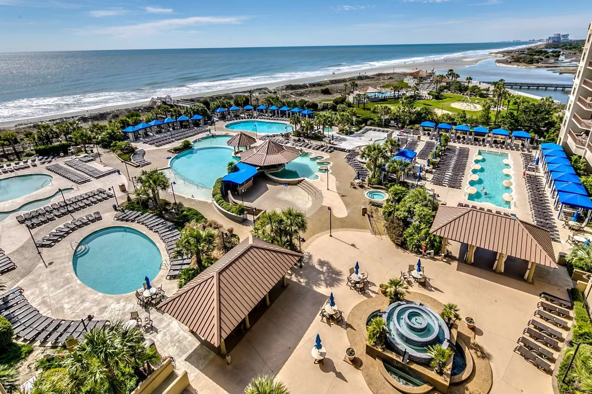 A beautifully landscaped outdoor pool area with multiple swimming pools, beachfront access, and poolside cabanas. Palm trees and lush gardens surround the space, offering both sun and shade for guests. The tranquil environment invites relaxation, while the scene hints at both privacy and communal enjoyment.