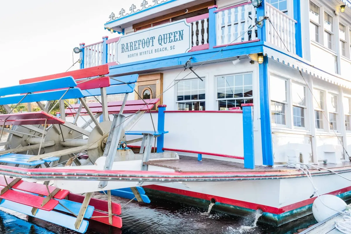 The image displays a charming, brightly-colored paddlewheel riverboat docked at the water’s edge, with clear signage and outdoor seating. The festive vibe suggests opportunities for unique dining and sightseeing experiences on the water.