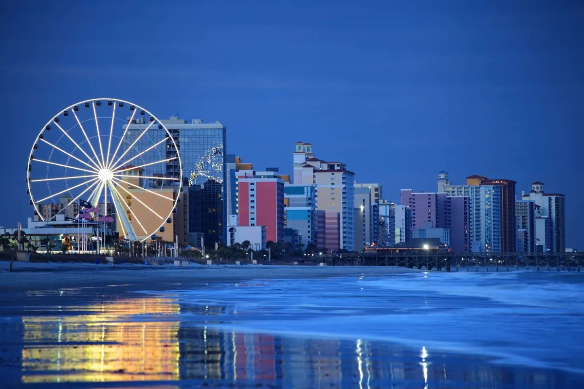 A lively nighttime coastal scene featuring a lit-up ferris wheel, colorful waterfront buildings, and sparkling reflections on the ocean. The festive atmosphere hints at nearby attractions and beachside adventures, with opportunities for amusement rides and waterfront dining.