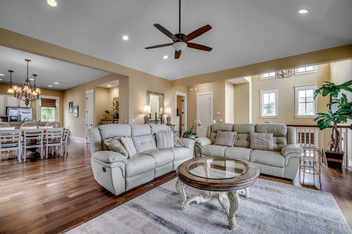 A bright, open-concept interior featuring leather sofas in the living area, a sleek coffee table, chandeliers, and a visible dining area beyond. Large windows allow abundant sunlight to stream in, emphasizing the spacious layout and inviting atmosphere perfect for gatherings and celebrations.