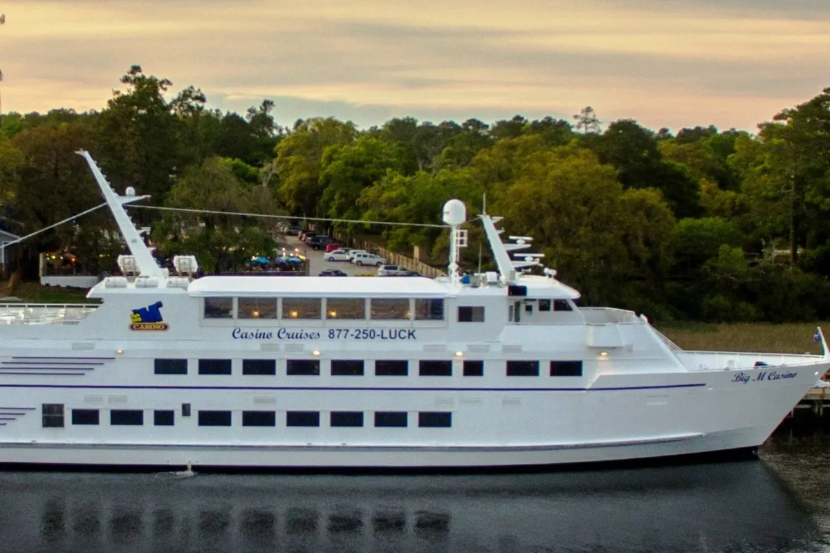 A large white cruise ship with blue accents is docked, with visible signage advertising casino cruises. The deck offers areas for casual dining and socializing, hinting at entertainment and nighttime activities. Sunset lighting enhances the sense of adventure and excitement.