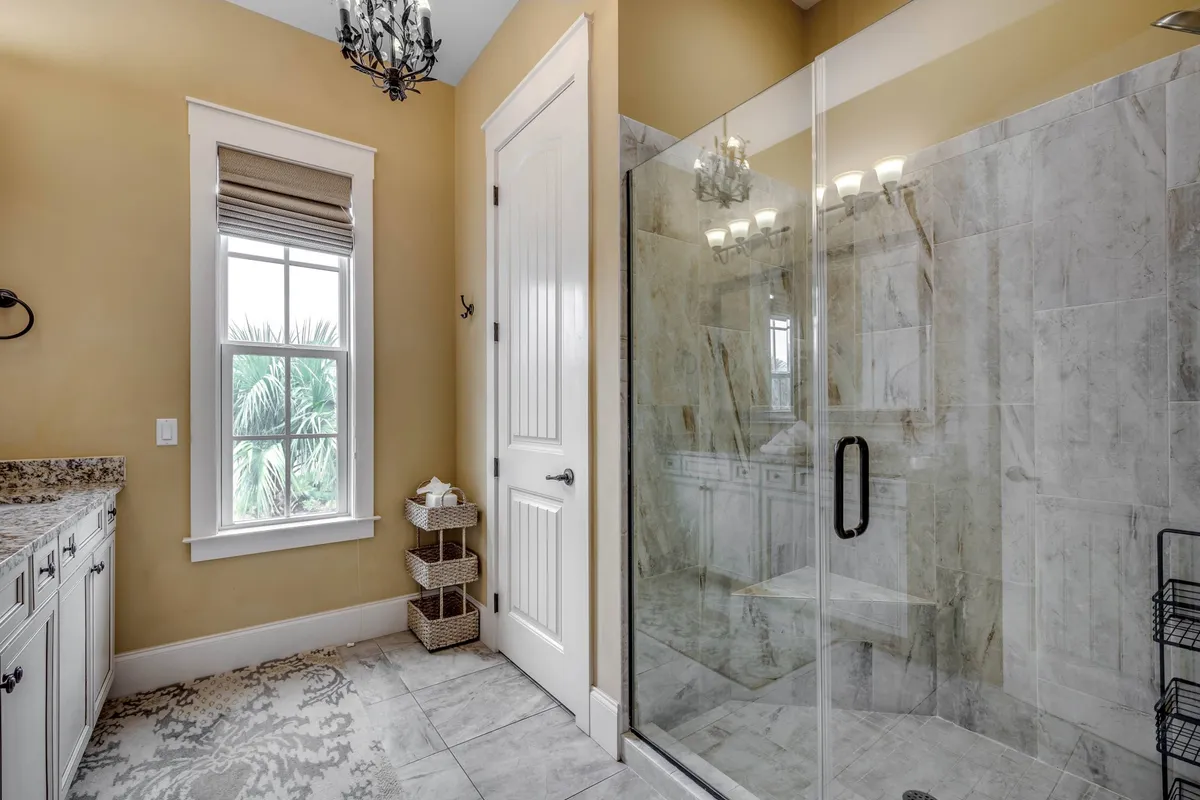 A stylish and elegant bathroom featuring a glass shower enclosure, dual sinks, a decorative chandelier, and a window with palm views. The space feels tranquil, perfect for relaxing baths or morning routines.