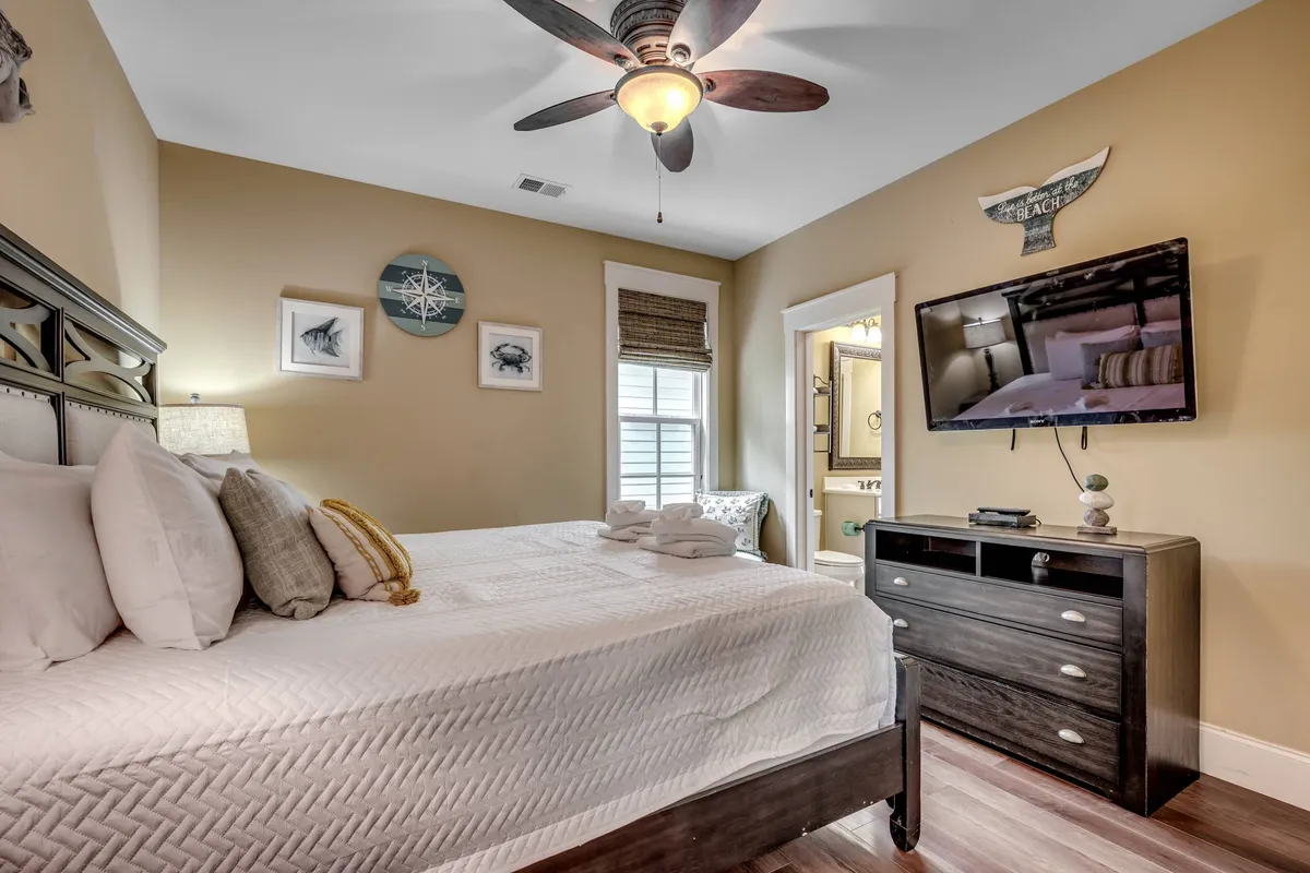 A well-appointed bedroom with a bed topped by decorative pillows, a wall-mounted television, ceiling fan, and framed artwork. Natural light streams in, highlighting the inviting atmosphere and comfortable bedding.