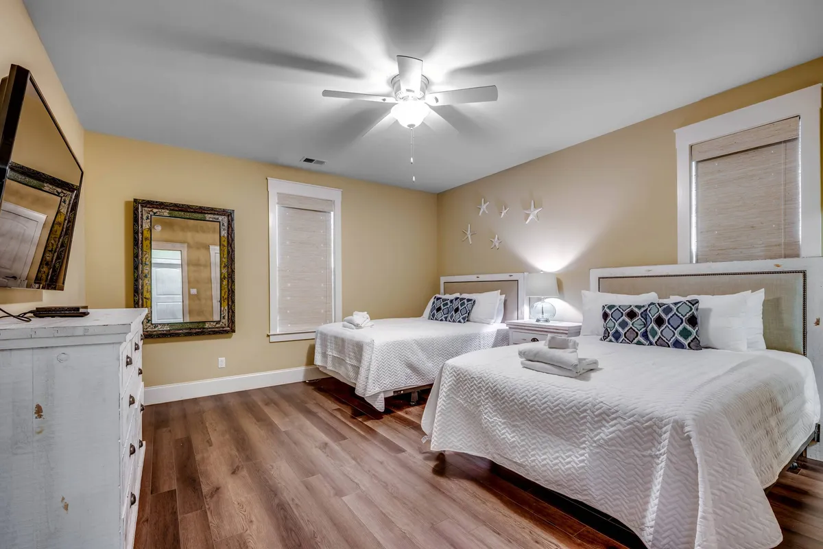 A calm, airy bedroom with two beds, decorative mirror, wall decor with stars, a dresser, and a television. The space is bright with abundant natural light, ideal for families or groups sharing the space.