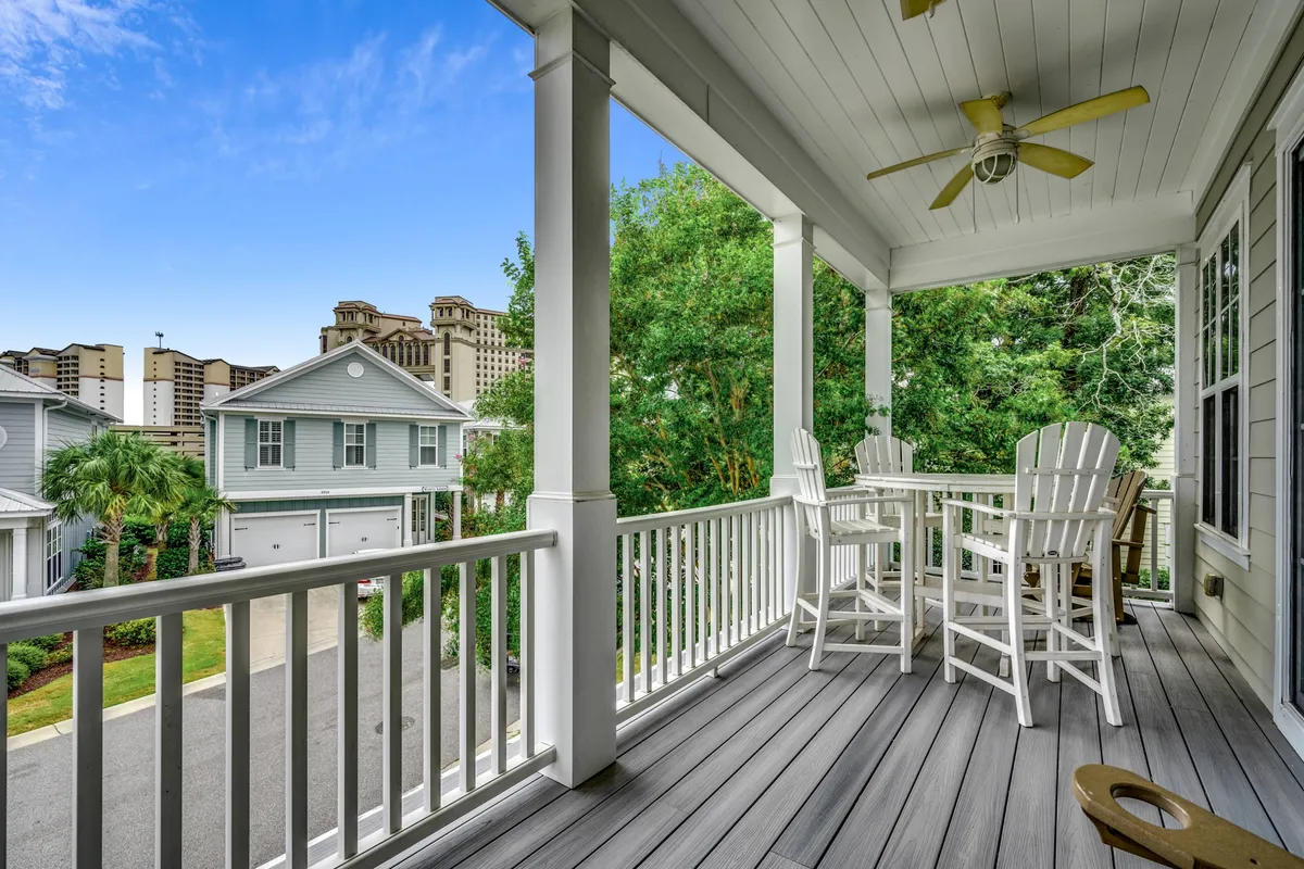 A breezy wooden deck with a bar-height table and chairs is shaded by a ceiling fan, surrounded by greenery and offering glimpses of a city skyline. The scene suggests relaxed conversation, open-air dining, and comfort on even warm days.