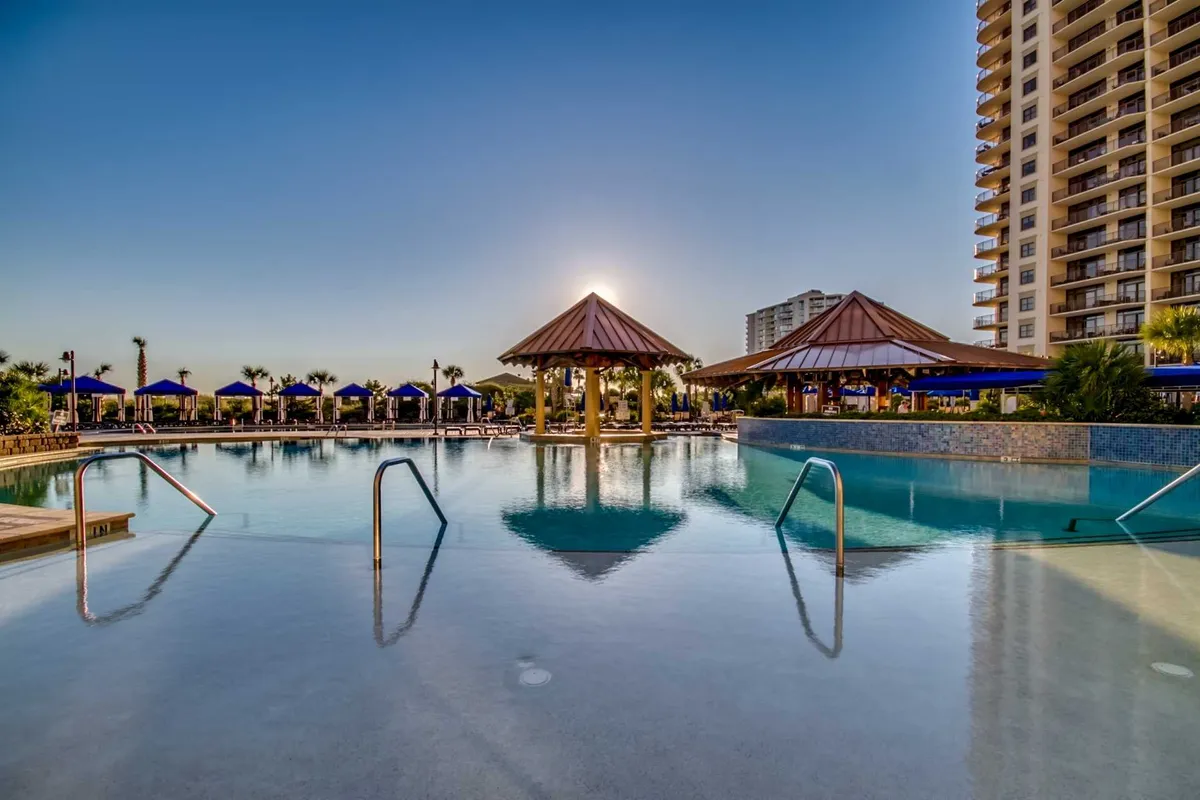 A contemporary pool area with sparkling water, thatched-roof cabanas, and a modern resort building. The sun sets behind the scene, casting a golden glow on the area and creating an inviting, luxurious atmosphere ideal for unwinding and social interaction.